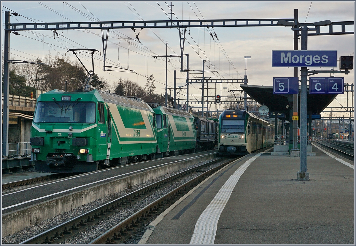 Die beiden BAM MBC Ge 4/4 22 und 21  schieben ihren bereits umfahrenen Güterzug mit 28 Achsen auf die Rollbockanlage, wo die Normalspurgüterwagen de BAM /MBC wieder ihr Normalspurgleis erreichte, Der Zug wird später mit der BAM MBC Re 4/4 II nach Gland gefahren. Rechts im Bild wartet ein BAM MBC Be 4/4  SURF auf die Abfahrt nach Bière.
Ganz rechts im Bild aber kaum erkennbar wartet der Gegenzug auf den Spur und Fahrrichtungswechsel; auf jedenfall ergibt die Kreuzung der beiden Züge eine  Kaisermanöver  vom Feinsten. 
22. Feb. 2017