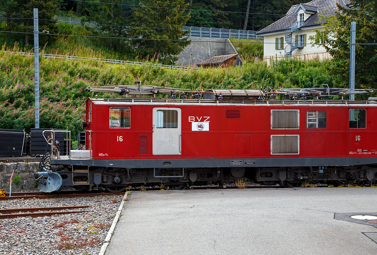 Leider nicht ganz erwischt, der Rest war aber auch vom Stationsgebäude verdeckt....
Die BVZ HGe 4/4 I - 16 der ehem. Brig-Visp-Zermatt-Bahn (damals noch VZ - Visp–Zermatt-Bahn, heute MGB - Matterhorn-Gotthard-Bahn) ist am 07.09.2021 beim Bahnhof Hospental abgestellt. 

Die HGe 4/4 – 16 ist eine schmalspurige (1.000 mm) gemischte Zahnrad- und Adhäsions-Elektrolokomotive der Visp-Zermatt-Bahn (VZ), die 1939 von SLM (Schweizerische Lokomotiv- und Maschinenfabrik) in Winterthur gebaut wurde, der elektrische Teil ist von der MFO (Maschinenfabrik Oerlikon). Diese Lok war später das Vorbild für die etwas stärkeren FO HGe I 31–37 der ehemalige Furka-Oberalp-Bahn (FO).

Die BVZ16, die nach einem Umbau für Pendelzugbetrieb 1987 als Reserve für die Pendelzüge Täsch–Zermatt eingesetzt wurde, kam nach der Übernahme des Pendelzugverkehrs durch die Niederflur-Panoramatriebzüge BDSeh 4/8 in den Jahren 2003 und 2005 vor Dienstzügen zum Einsatz. Sie wurde im Sommer 2007 mit den Scherenstromabnehmern von Lokomotive Nummer 37 versehen, die Nummer 16 besaß zwischenzeitlich zwei Einholmstromabnehmer, zu einem symbolischen Preis an die Dampfbahn Furka-Bergstrecke (DFB) verkauft. Sie wurde 2007 in Gletsch abgestellt. 2018 wurde das Fahrzeug in den Bestand von MGBahn-Historic aufgenommen. Nun ist sie vorübergehend in Hospental abgestellt, eine Aufarbeitung in der MGB-Werkstätte im Glisergrund ist in der Planungsphase.

Technik:
Die VZ-Lokomotive 16 und später die FO-Lokomotiven 31–37 sind eine Weiterentwicklung der VZ HGe 4/4 11–15, erhielten jedoch einen neu konstruierten Lokomotivkasten in Stahlleichtbauweise, weil der Aluminiumkasten der VZ HGe 4/4 11–15 anfänglich nicht befriedigte. Zur Verringerung des dadurch bedingten Mehrgewichts wurden unter anderem die Vorbauten weggelassen und die gesamte technische Ausrüstung in den dazu verlängerten Innenraum des Lokomotivkastens verlegt. Statt der Vorbauten entstanden an den Enden offene Plattformen, wie dies damals bei verschiedenen Elektrolokomotiven der Fall war, Beispielsweise an den Be 6/8 der damaligen Berner Alpenbahn-Gesellschaft Bern-Lötschberg-Simplon (BLS), die ab dem Jahre 1926 abgeliefert wurden. Über diese Plattformen konnten die Führerstände betreten werden und erhielten damit die charakteristische, nicht symmetrische Fahrzeugfront. Die Transformatoren bekamen wieder eine Ölkühlung.

Die ursprünglichen FO-Lokomotiven haben zudem einen Anschluss für die Speisung der 1941, 1943 und 1945 von SLM/MFO gelieferten elektrischen Schneeschleudern Xrot e 4931–4933.

Die Lokomotiven der FO, wie auch der VZ, waren nicht für die Zahnradbahnstrecke Andermatt–Göschenen der damaligen Schöllenenbahn (SchB) ausgelegt, die 1961 in die damalige FO integriert wurde.

Drehgestelle:
Die Lokomotiven besitzen zwei zweiachsige Drehgestelle mit einem kurzen Radstand und einem kombinierten Antrieb. Die beiden außerhalb der beiden Triebachsen liegenden Motoren arbeiten gleichzeitig und untrennbar auf die jeweilige Antriebsachse und dessen Triebzahnrad. Die Zug- und Stoßvorrichtungen sind jeweils direkt mit dem Drehgestellrahmen verbunden. Im Unterschied dazu werden diese Vorrichtungen heute bei Drehgestelllokomotiven in der Regel am Lokomotivkasten befestigt. Die bei der Beförderung von Zügen auftretenden Zug- und Druckspannungen wirken im Normalbetrieb im Falle dieser Lokomotiven, da diese in der Regel an der Zugspitze eingesetzt wurden, zunächst direkt auf das in Fahrtrichtung hintere Drehgestell. Erst über eine zentrale kugelige Drehpfanne werden die Kräfte auf eine Brücke, die aus kräftigen Profilen zusammengenietet ist, und von dort auf das vordere Drehgestell übertragen. Auf dieser Brücke aufgebaut ist der leicht konstruierte Lokomotivkasten.

TECHNISCHE DATEN (die der FO HGe I 31–37 weichen etwas ab):
Spurweite: 1.000 mm
Achsformel: Bozz’Bozz’
Hersteller: SLM Winterthur / Maschinenfabrik Oerlikon
Dienstgewicht: 46,6 t
Länge über Puffer: 14.100 mm
Drehzapfenabstand: 6.710 mm
Achsabstand im Drehgestell: 2.010 mm
Höchstgeschwindigkeit : 50 km/h (Adhäsion)  / 25 km/h (Zahnrad)
Anzahl der Motoren: 4
Stundenleistung: 736 kW (1.000 PS)
Stromsystem: Einphasenwechselstrom 11 kV / 16,7 Hz,
Zahnstangensystem : Systems Abt (mit zwei Lamellen)
