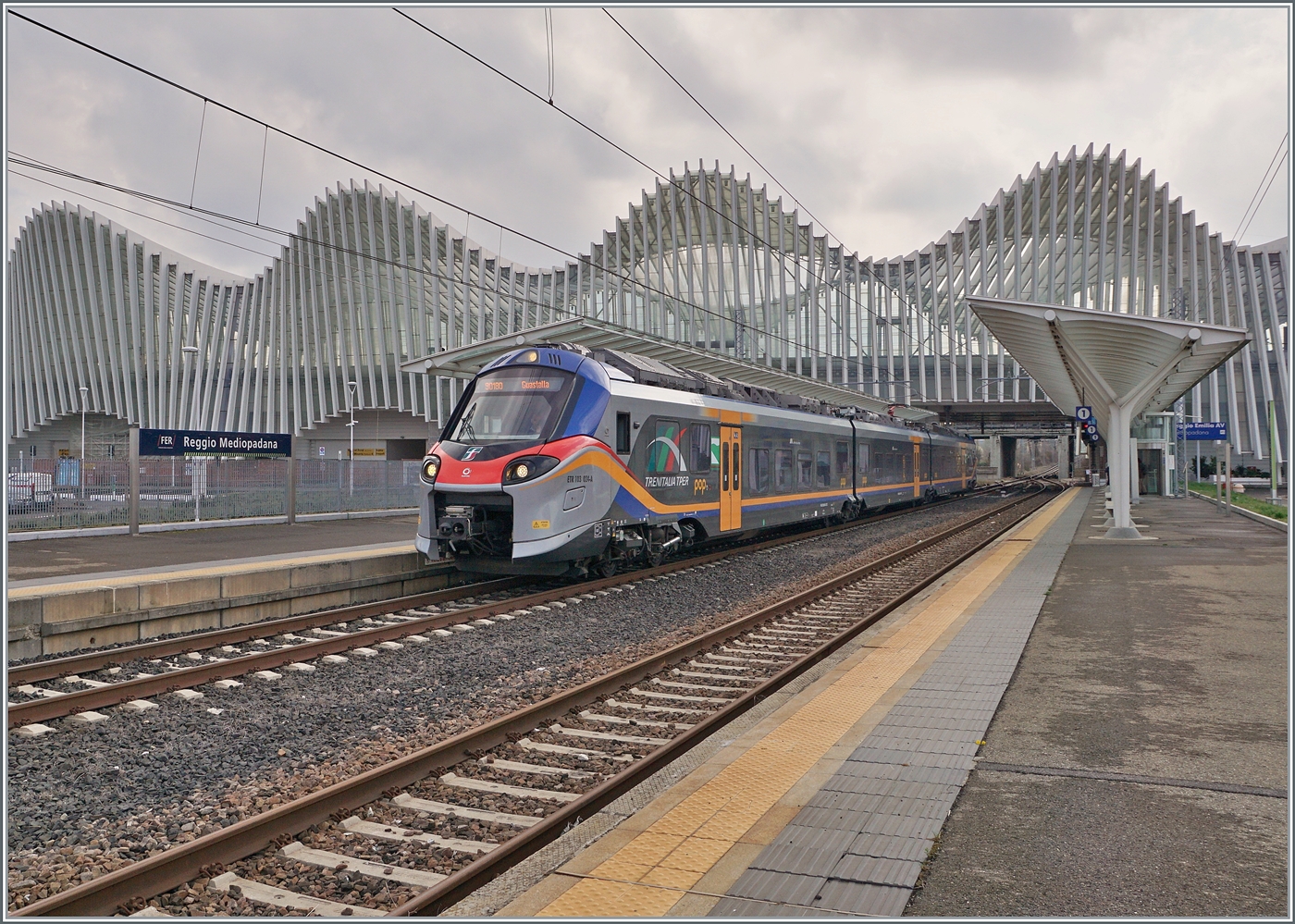 Der FS Treniatalia ETR 103-024 der Tochtergesellschaft TPER (Trasporto Passeggeri Emilia Romagna) ist als Regionalzug 90180 von Reggio Emilia nach Guastalla unterwegs und verlässt den Bahnhof Reggio Mediopadana. Im Hintergrund ist der imposante Bahnhof von Reggio Emilia AV zu erkennen.

14. März 2023