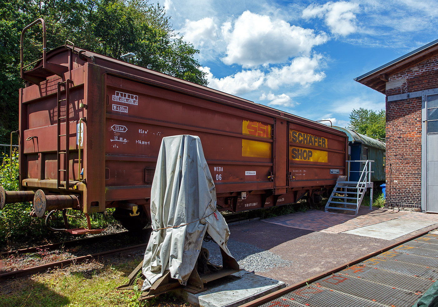 Der gedeckte zweiachsige Schiebewand-/Schiebedachwagen WEBA 06 - 10 80 0715 000-7 D-WEBA (ex 10 80 0715 000-7 D-DB) der Gattung Tbis 869 - Bauform A (für Büromöbel der SSI Schaefer Shop GmbH) ehemals der WEBA (Westerwaldbahn des Kreises Altenkirchen GmbH), ausgestellt am 07 Juli 2024 beim Erlebnisbahnhof Westerwald der Westerwälder Eisenbahnfreunde 44 508 e. V. in Westerburg, hier war Lokschuppenfest.

Beschreibung:
Die Beschaffung erster Hubschiebedachwagen begann bereits 1949. Die Wagen verfügten über ein zweiteiliges Dach, von denen ein Teil über das andere geschoben werden konnte. In der Mitte des Wagens war ein der Dachform angepasstes Querjoch angebracht, um einerseits die Spur der Dachlaufwerke halten zu können, andererseits das Dach dicht verschließen zu können. Betätigt wurde das Dach durch ein an den Kopfendenden angebrachte hoch liegende Bühne, die über Leitern erreicht wurden.

Ab 1966 wurden von der DB bei der Waggonunion Siegen (eigentlich Dreis-Tiefenbach, zuvor Siegener Eisenbahnbedarf A.G.) die ersten Wagen der Gattung Tbis 869 der Bauform A beschafft. Bei dieser Bauform konnte das Dach auch nur von einer kleinen oben liegenden Bühne an den Stirnwänden geöffnet werden. Zwei Jahre später wurde die Bauform B dieser Gattung gefertigt. Hierbei war es nun möglich das Dach mittels Handräder an einer Stirnseite der Waggons zu öffnen. Es war nun nicht mehr nötig auf das Dach zu steigen und verminderte somit auch die Unfallgefahr.

Eingesetzt wurden diese Waggons für den Transport von großen nässeempfindlichen Stückgütern (bei diesem Wagen waren es später Büromöbel), durch das öffnungsfähige Dach könnte die Ladung auch mittels Kranen schnell be- und entladen werden. Von den ca. 2.000 beschafften Fahrzeugen gelangten noch gut 1.400 zur DB AG, dort wurden sie aber zügig ausgemustert und durch neuere Bauarten ersetzt. 

TECHNISCHE DATEN:
Spurweite: 1.435 mm (Normalspur)
Anzahl der Achsen: 2
Länge über Puffer :14.000 mm
Achsabstand: 8.000 mm
Eigengewicht: 18.400 kg
Ladelänge:12.800 mm
Ladefläche: 33,0 m²
Rauminhalt: 85,0 m³
Seitenwand-Türöffnung (B × H): 5.660 × 2.100 mm
Lichte Öffnung bei geöffnetem Dach: 6.000 × 2.530 mm
Fußbodenhöhe über SO: 1.200 mm
Höchstgeschwindigkeit: 100  km/h 
Maximales Ladegewicht: 18,5 t (Streckenklasse C oder höher)
Kleinster bef. Gleisbogenradius:  35 m
Bremse: Frein O-GP 
Intern. Verwendungsfähigkeit: RIV (ursprünglich)
