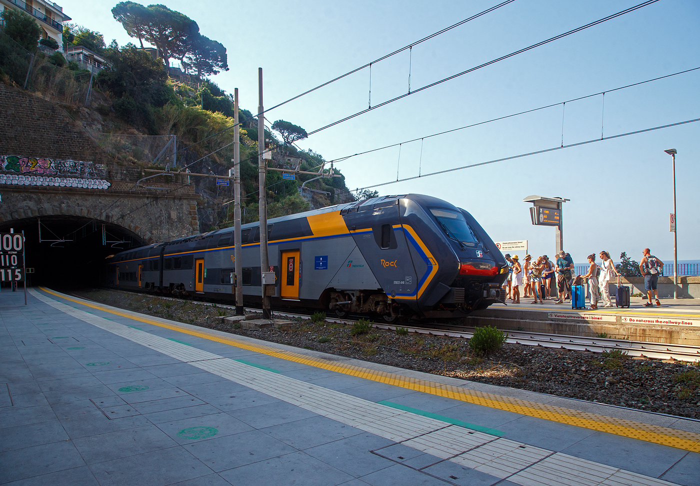 Der Trenitalia “Rock” ETR 521-048, ein fünfteiliger Elektrotriebzug vom Typ Hitachi Caravaggio, erreicht am 22.07.2022 den Bahnhof Riomaggiore (Cinque Terre). Er fährt als Cinque Terre Express (Regionale) von La Spezia Levanto, via Riomaggiore, Manarola, Corniglia, Vernazza, Monterosso, nach Levanto.

Die Züge des Cinque Terre Express, obwohl sie alle ¼ Stunde fahren, sind immer sehr gut gefüllt. Nicht immer findet man einen Sitzplatz. Für die Fahrt mit den Cinque Terre Express muss man ein spezieles Ticket lösen, z.B. Trenitalia nationale Tageskarten gelten hier nicht. 

Wer wie wir die Cinque Terre Card Treno (Zugkarte) besitzt, kann dann auch mit den Minibusen des Cinque Terre Parks (im Preis der Cinque Terre Treno Card eingeschlossen) in den fünf Dörfern kostenlos fahren. Zudem hat man mit dieser Karte auch Zutritt zu den beiden gebührenpflichtigen Wanderwegen des Cinque Terre Parks.