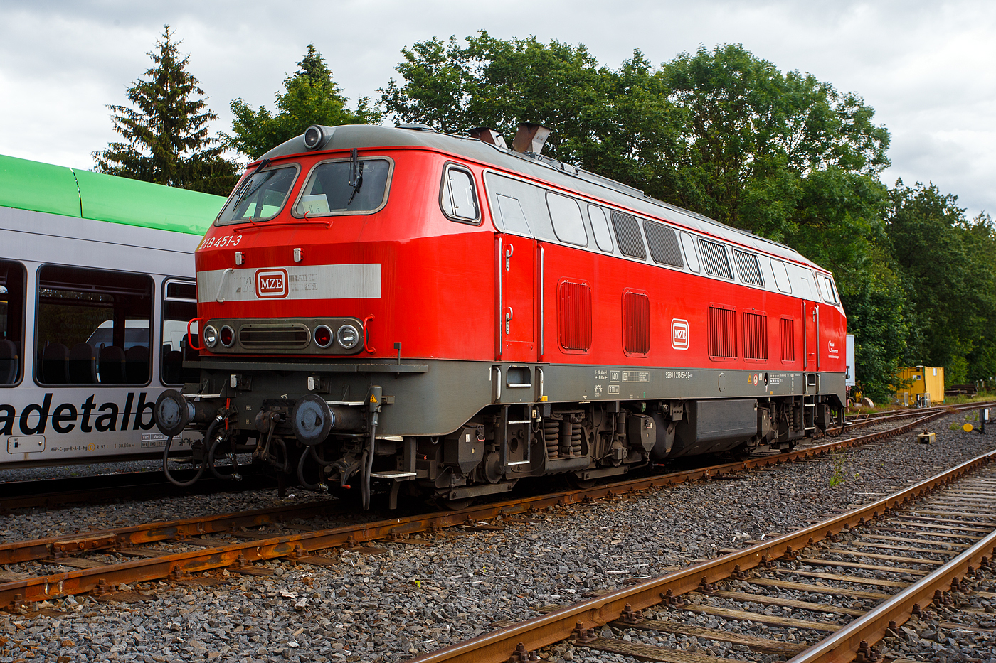 Die 218 451-3 (92 80 1218 451-3D-MZE) der MZE - Manuel Zimmermann Eisenbahndienstleistungen, Hellenhahn-Schellenberg (Ww), ist am 16 Juni 2024 auf dem Betriebshof der WEBA (Westerwaldbahn des Kreises Altenkirchen GmbH) auf der Bindweide bei Steinebach/Sieg abgestellt.

Die V 164 wurde 1978 bei Henschel in Kassel unter der Fabriknummer 32045 gebaut und als 218 451-3 an die DB geliefert. Bis April 2016 fuhr sie für die DB Regio AG – Nord als 92 80 1218 451-3 D-DB im Regionalbahn Einsatz in Niedersachsen und Bremen. Im Jahr 2016 ging sie dann an die LWC Lappwaldbahn Cargo GmbH in Braunschweig und war Ende 2021 an die Hering Bau GmbH & Co. KG in Burbach vermietet. Im Juni 2023 wurde sie dann an die MZE - Manuel Zimmermann Eisenbahndienstleistungen verkauft.

Die Baureihe 218 ist das zuletzt entwickelte Mitglied der V 160-Lokfamilie. Viele Gemeinsamkeiten der Baureihen V 160 bis V 169 (spätere 215 bis 219) wurden in ihr zusammengefasst. Bei der Baureihe 218 wurde von der Baureihe 217 die elektrische Zugheizung übernommen, von den Prototypen der Baureihe 215 übernahm man den 1.840-kW-Motor (2.500 PS), wobei ein Hilfsdieselmotor zum Betrieb des Heizgenerators überflüssig wurde. Später wurden noch stärkere Motoren mit 2.700 PS (KHD Lizenzbau Pielstick16 PA4 V 200) oder mit 2.800 PS (MTU 12 V 956 TB 11) verbaut. Den verbauten Motor kann man anhand der Anordnung der Abgashutzen auf dem Dach erkennen.

Im Jahr 1966 bestellte die Deutsche Bundesbahn zunächst zwölf Vorserien-Lokomotiven der Baureihe V164. Die ersten Lokomotiven wurden ab 1968 von der Firma Krupp ausgeliefert. Von der Deutschen Bundesbahn wurden sie aber als Baureihe 218 in Dienst gestellt. Die Serienbeschaffung (unter Beteiligung von Henschel, Krauss-Maffei und MaK in Kiel) erfolgte von 1971 bis 1979 mit 398 weiteren Maschinen. Hinzu kam 1975 nach einem Unfall die 215 112, die nach ihrer Instandsetzung zur 218 399 wurde. Die Auslieferung erfolgte in vier Bauserien (218 101-170, 171-298, 299-398, 400-499), bei denen es geringe Veränderungen gab.

Die 140 km/h schnellen und 2.500 bis 2.800 PS starken ''BB-Loks“  wurden im Reise - und Güterzugdienst eingesetzt. Die elektrische Zugheizung und die Wendezug- und Doppeltraktionssteuerung machen die Baureihe 218 zu einer universal verwendbaren Lok. Die Baureihe 218 bewährte sich im Betriebsdienst und galt noch bis ins Jahr 2000 als die wichtigste Streckendiesellok der Deutschen Bahn AG. Leider wurden immer mehr Leistungen im Personennahverkehr durch Triebwagen ersetzt. Dadurch und durch Betreiberwechsel waren immer mehr Lokomotiven der Baureihe 218 bei der Deutschen Bahn AG überflüssig geworden. Im Juli 2009 waren noch etwa 200 Exemplare im Einsatz. Eine echte Nachfolgerin für die Baureihe 218 ist noch nicht in Sicht.

TECHNISCHE DATEN:
Spurweite: 1.435 mm (Normalspur)
Achsformel:  B'B'
Spurweite:  1.435 mm
Länge: 16.400 mm
Breite des Lokkastens: 3.136 mm
Höhe des Lokkastens: 4.275 mm über Schienenoberkante
Drehzapfenabstand:  8.600 mm
Achsabstand im Drehgestell: 2.800 mm
Treibraddurchmesser: 1 000 mm (neu) / 920 mm (abgefahren)
Gewicht:  79 Tonnen
Achslast: 19,5 t
Motor: MTU wassergekühlter V12-Zylinder-Viertakt -Dieselmotor vom Typ 12 V 956 TB 11 (abgasoptimiert ) mit Direkteinspritzung, Abgasturboaufladung und Ladeluftkühlung
Motorleistung: 2.800 PS (2.060 kW) bei 1,500 U/min
Motor-Hubraum: 114,7 Liter
Anfahrzugkraft: 235kN 
Dauerzugkraft: 175kN
Höchstgeschwindigkeit:  140 km/h (im Langsamgang 100 km/h)
Motorleerlaufdrehzahl (ohne Zugheizung) : 630 - 650 U/min 
Motorleerlaufdrehzahl (mit eingeschl. Zugheizung): ca 1000 U/min 
Kleinste Dauergeschwindigkeit (im Schnellgang): ca. 28 km/h
Kleinste Dauergeschwindigkeit (im Langsamgang) ca. 18 km/h
Getriebe: MTU-Getriebe K 252 SUBB (mit 2 hydraulische Drehmomentwandler)
Leistungsübertragung: hydraulisch
Kleinster befahrbarer Gleisbogen: R = 100 m
Tankinhalt:  3.150 l
Sandvorrat: 400kg
Bremse: KE-GPP2R-H mZ
Bemerkung: Den verbauten Motortyp kann man auch anhand der Anordnung der Abgashutzen auf dem Dach erkennen.

Aufbau und Ausrüstung
Motor und Getriebe sind elastisch im Rahmen gelagert. Der Rahmen stützt sich über je vier Schraubenfedern pro Drehgestell und Seite auf die Drehgestelle ab. Ab 218 299 wurden dabei Flexicoil-Federn verwendet, die eine gleitstücklose Bewegung der Drehgestelle ermöglichen (Atlas-MaK-Drehgestelle). Zug- und Bremskräfte werden über tiefliegende Querträger mit Drehzapfen übertragen. Die Aufbauten sind in Schalenbauweise aus Blechen und Profilstahl gefertigt. Zwischen den beiden Endführerständen verlaufen zwei Seitengänge.

Der Antrieb erfolgt über vier sehr lange Gelenkwellen von einem zentral liegenden Strömungsgetriebe mit Wendegetriebe. Dieses Getriebe ist mit zwei Drehmomentwandlern ausgerüstet. Die Schaltung von Wandler I auf Wandler II erfolgt automatisch und ohne Zugkraftunterbrechung. Außerdem ist das Getriebe mit zwei Geschwindigkeitsstufen ausgerüstet. Die kleine Geschwindigkeitsstufe (Langsamgang) ermöglicht höhere Zugkräfte, allerdings eine niedrigere Endgeschwindigkeit von 100 km/h, die große Geschwindigkeitsstufe (Schnellgang) erlaubt die höhere Höchstgeschwindigkeit von 140 km/h, jedoch mit niedrigeren Zugkräften. Die Umschaltung von Langsam- auf Schnellgang erfolgt ausschließlich bei stehender Lokomotive. Zudem sind die Serienlokomotiven mit einer verschleißfreien hydraulischen Bremse ausgerüstet.

Im Laufe der Zeit wurden verschiedene Dieselmotoren als Antrieb eingesetzt: Die ersten Serien erhielten den MTU MA 12V 956 TB10 mit 2.500 PS. Um den bei diesen Motoren eintretenden Zugkrafteinbruch bei voller Heizleistung zu umgehen, wurden bei späteren Maschinen einige von KHD in Lizenz gebaute französische Pielstick 16 PA4 V200 mit rund 2,700 PS eingebaut. MTU zog nach und entwickelte aus dem TB10 den 2,800 PS leistenden stärkeren TB11, der in weit mehr als der Hälfte aller 218 zum Einbau kam. Durch schlechte Abgaswerte und fortschreitenden Verschleiß der Motoren sah sich der Betreiber in den letzten Jahren zu einem umfassenden Modernisierungsprogramm gezwungen. Zunächst wurden etliche TB11-Motoren mit einem Umrüstsatz abgasoptimiert. Weiterhin wurden mehrere diesem Stand entsprechende TB11-Motoren neubeschafft, die zudem über ein elektronisches Motormanagement verfügen. Parallel wurden auch gänzlich neue Motoren erprobt, was letzten Endes zur Beschaffung zahlreicher 16V 4000 R40/R41 von MTU führte. Außerdem wurden in drei Lokomotiven (218 329, 330 und 339) Motoren der Firma Caterpillar eingebaut. Durch den Zuwachs an Motoren wurde die Bestückung der Lokomotiven kräftig durchmischt. 

Ab 1981 wurden die Lokomotiven mit zusätzlichen Abgashutzen auf dem Dach ausgerüstet, um die Abgase an der Fahrleitung vorbeizuleiten und so deren Verschmutzung zu verringern, die durch Abgasklappen in den Hutzen erhöhte Austrittsgeschwindigkeit sollte zudem die Belästigung der Fahrgäste durch Abgase verringern. Die Anordnung der Hutzen ist ein Hinweis auf den in der Lokomotive eingebauten Motor: Lokomotiven mit Zwölfzylindermotor (TB10/TB11) haben entsprechend der einseitig am Motor angebrachten Abgasanlage nebeneinander angeordnete Hutzen, bei Sechzehnzylindermotoren (Pielstick, MTU 4000) mit Abgasanlagen an beiden Motorseiten sind die Hutzen versetzt angebracht.
