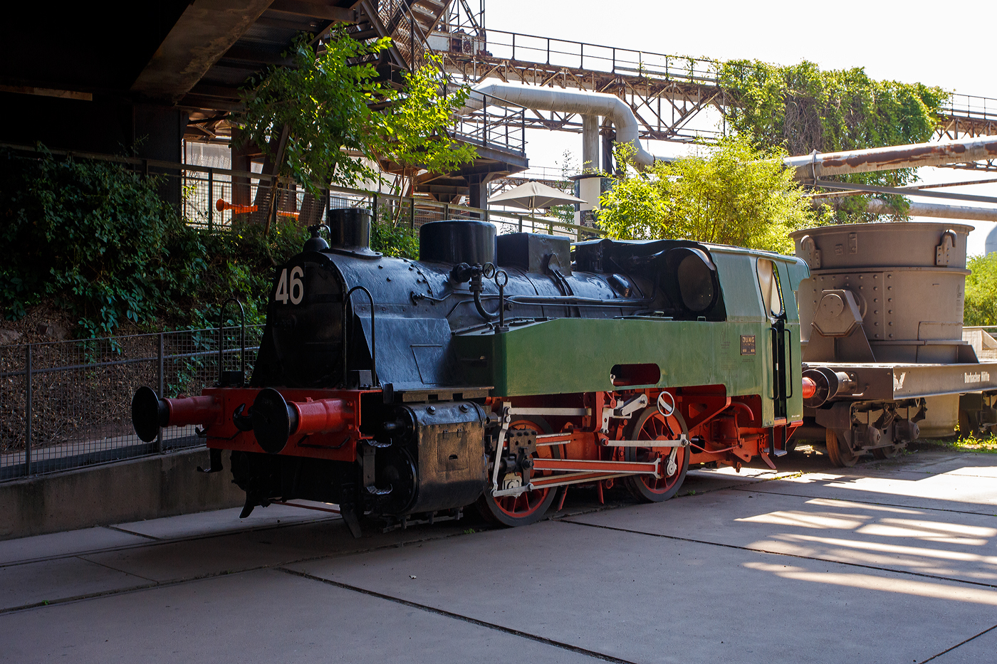 Die Normalspur-Dampflokomotive 46 der V�lklinger H�tte (ehem. R�chling'schen Eisen- und Stahlwerke GmbH in V�lklingen/Saar, ab 1982 ARBED-Saarstahl GmbH, ab 1986 Saarstahl V�lklingen GmbH), eine Jung Hochofenlok niedriger Bauart, abgestellt mit einem offenen Roheisenpfannenwagen der Burbacher H�tte, hier am 01 Juli 2025 am Gel�nde V�lklinger H�tte als Denkmal. Heutiger Eigent�mer ist die Initiative V�lklinger H�tte e.V..

1964 wurde die Lok Nr. 46 mit der Fabriknummer 13835 im Auftrag der R�chling'schen Eisen- und Stahlwerke als letzte rostgefeuerte Dampflok der Firma Arnold Jung, Lokomotivfabrik GmbH, in Jungenthal bei Kirchen an der Sieg gebaut und am 09.12.1964 geliefert. Einsatzgebiet war der Roheisentransport von den sechs Hoch�fen zum Thomasstahlwerk, wobei die besonders beengten Verh�ltnisse auf der V�lklinger H�tte eine besondere Bauform erforderten. Die Verbindung zwischen Eisenwerk und Stahlwerk war nur durch einen Tunnel m�glich, da die Deutsche Bahn durch ihre Gleisanlagen die H�tte in zwei Bereiche teilt. Dieser Tunnel, der Roheisenkanal, ist so niedrig, dass die zweiachsige Lok nur eine H�he von 2.640 mm �ber der Schienenoberkante und eine Breite von 2.450 mm aufweist. Die Loks 44 (11945/Baujahr 1953) und 45 (13240/Baujahr 1959), die nach vorliegenden Unterlagen technisch baugleich sind, wurden schon vorher (von der Arnold Jung GmbH an die V�lklinger H�tte geliefert. 

Um speziell die F�hrerhausr�ckwand, die Lampen, aber auch die Kupplungselemente zwischen Lok und dem ersten Wagen vor fl�ssigen gl�henden Spritzern von herabfallenden Roheisen zu sch�tzen, erhielten die Lokomotiven ein nach hinten verl�ngertes Dach. Die Puffer wurden im Stahlwerk den dort vorhanden Roheisenpfannenwagen angepasst. Alte Jung Mitarbeiter berichten, an der Lok h�tte man kaum was verdient, weil l�nst nicht mehr Dampflokteile ab Lager vorhanden waren umd z.T. m�hsam und teuer beschafft werden musste.

Im Zuge der Modernisierung und Rationalisierung wurden die Roheisendampfloks 1981 nicht mehr ben�tigt. W�hrend die Lokomotiven 44 und 45 an ein Museum im Ruhrgebiet verkauft wurden, gelangte die Lokomotive 46 als technisches Denkmal 1982 zur Universit�t Kaiserslautern. Diese schenkte die Lokomotive 46 der Initiative V�lklinger H�tte, die sie mit Hilfe von Sponsoren am 9. Februar 1999 nach V�lklingen zur�ckholte.

TECHNISCHE DATEN:
Baujahre: 1953,1959 und 1964
Gebaute Anzahl: 3
Spurweite: 	1.435 mm (Normalspur)
Achsformel: B
L�nge: 7.600 mm
Achsabstand: 2.000 mm
Triebraddurchmesser: 1.000 mm (neu)
Breite: 2.450 mm
H�he: 2.650 mm
Dienstgewicht: 26 t
H�chstgeschwindigkeit: k. A. 
Leistung: 250 PS
Zugkraft: 7,5 t
Zylinder: 2 St�ck, � 380 mm x 500 mm Hub
Dampfdruck: 14 bar
Heizfl�che: 58,92 m�
Rostfl�che: 1,17 m�
Anzahl Siederohre: 178 St�ck
Durchmesser Siederohre: 33/38 mm
L�nge Siederohre: 2.540 mm
Wasservorrat: 2.500 l (2,5 m�)
Kohlenvorrat: 500 kg