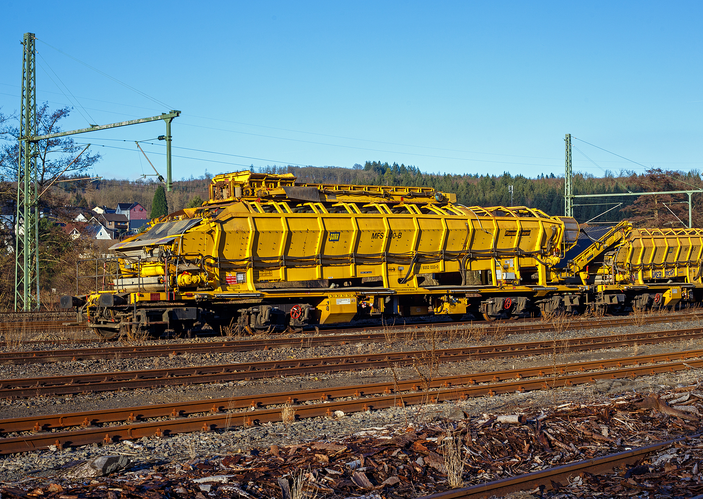 Drei Plasser & Theurer Materialf�rder- und Siloeinheiten (MFS 100) der �sterreichischen Gleisbaufirme PORR AG (Wien) sind, mit einem Schutzwagen, am 26 Dezember 2024, in Scheuerfeld/Sieg abgestellt.

Hier im Vordergrund die P&T Materialf�rder- und Siloeinheit MFS 100 B mit integrierter Beladestation (in Transportstellungen oben auf dem MSF) 99 81 9352 033-0 A-PTU.

TECHNISCHE DATEN:
Hersteller: Plasser & Theurer, Linz an der Donau (�sterreich)
Spurweite: 1.435 mm
L�nge �ber Puffer: 22.900 mm
Achsanzahl: 8 (in 2 Doppel-Drehgestellen)
Drehzapfenabstand der Doppeldrehgestelle: 15.100 mm
Drehzapfenabstand der Einzeldrehgestelle: 3.900 mm Achsabstand der Einzeldrehgestelle: 1.500 mm
Motorleistung: 165 kW (wassergek�hlter Deutz BF6M 1013 C Motor f�r Bandantrieb)
Eigengewicht: 67 t
Nutzlast: 77 t ab D4
Fahrgeschwindigkeit in Schleppfahrt leer, max.: 120 km/h
Fahrgeschwindigkeit in Schleppfahrt beladen, max.:100 km/h
Fassungsverm�gen: 68 m�
F�rderleistung (materialabh�ngig), max.: 700 – 800 m�/h
Entladezeit: ca. 5 min
Besonderheit: mit integrierter Beladestation