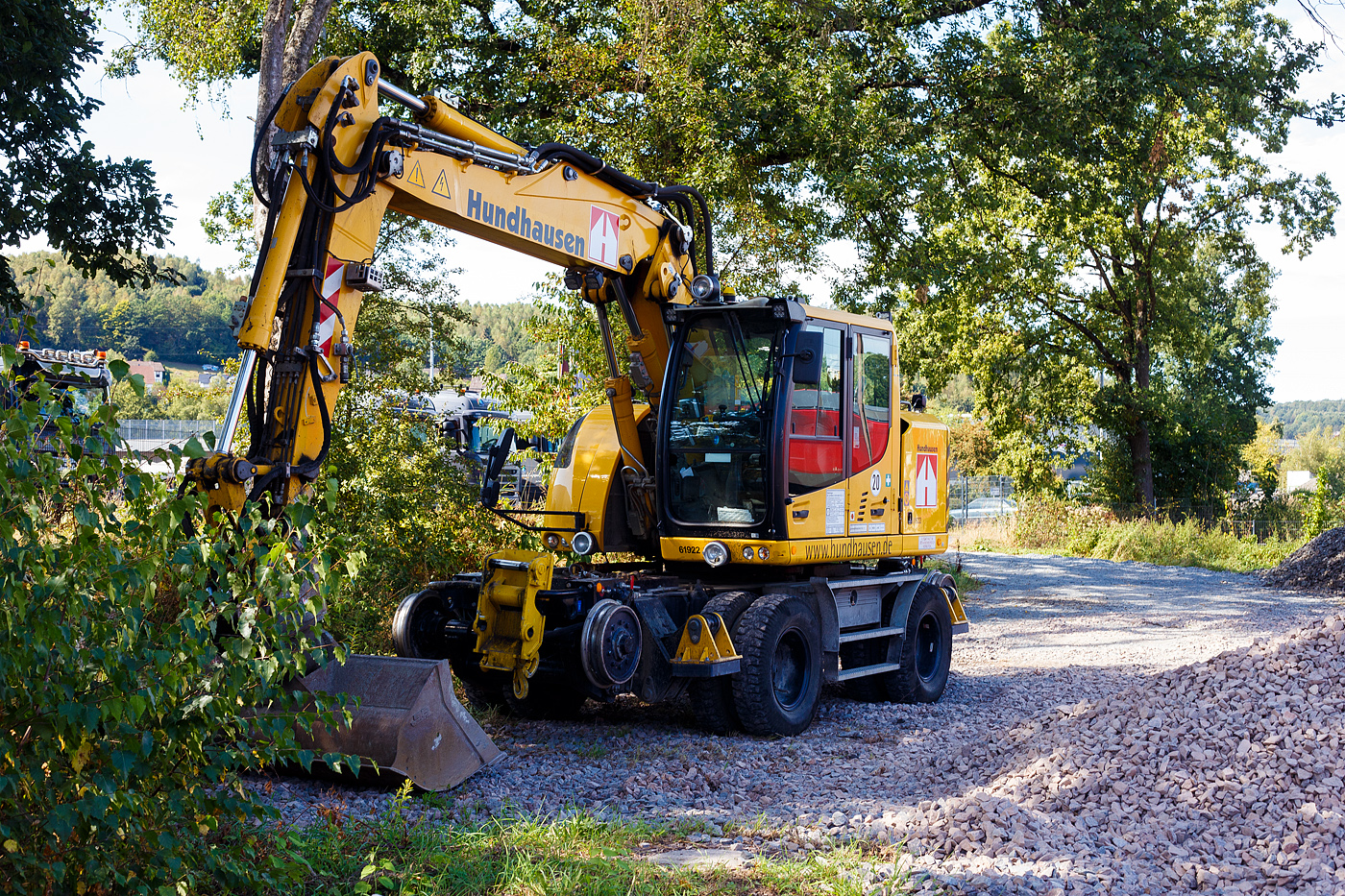 Liebherr Zweiwegebagger A 922 Rail Litronic mit Abstützpratzen, interne Nr. 61922 der Wilhelm Hundhausen GmbH & Co. KG, Siegen, eigestellt unter der Kleinwagen Nr. D-HHBAU 99 80 9903 752-0 abgestellt am 07 September 2024 in der Abstellgruppe in Kreuztal. 

Der A 922 Rail Litronic kann als Zweiwegemaschine wahlweise auf der Straße oder auf Eisenbahnschienen eingesetzt werden. An beiden Seiten des Unterwagens ist das Schienenfahrwerk angebracht. Während des Aufgleisens bringt das Schienenfahrwerk die Bereifung auf Schienenniveau, wobei die inneren Räder der Zwillingsbereifung den Fahrantrieb auf der Schiene übernehmen.

TECHNISCHE DATEN:
Spurweite: 1.435 mm (Normalspur)
Länge über Puffer: 6.400 mm
Achsabstand (Schiene): 5.700 mm
Laufraddurchmesser (Schiene): 500 mm (neu)
Eigengewicht: 23 t
Motorbauart: wassergekühlter 4Takt - 4-Zylinder-Reihendieselmotor mit Common-Rail-Einspritzsystem, Turbolader und Ladeluftkühlung
Motortyp: Liebherr D924 – FPT
Motorhubraum: 4,5 Liter
Motorleistung: 120 kW / 163 PS
Höchstgeschwindigkeit (Hg): 20 km/h (in Kreuzungen und Weichen 10 km/h)
Zugkraft: 117 kN
Bremse: Kfz.-/SFE-Bremse und Wagonbremsanlage
Zul. Anhängelast: 120 t (ungebremst 40 t)
Zur Mitfahrt zugel. Personenzahl: 1 (und Fahrer)
Antrieb auf Schiene: Über Reibantrieb der Straßenreifen
Hinterer Schwenkradius: 2.000 mm
