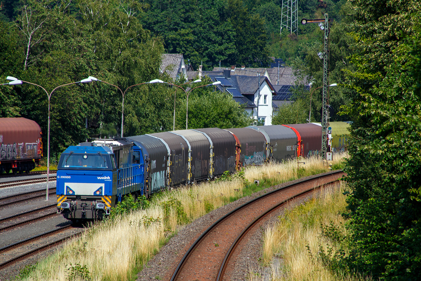 Teleblick auf die den lange Coilzug nachschiebende an die KSW vermietete 272 408-6 / D20 der Bentheimer Eisenbahn AG (92 80 1272 408-6 D-BE), ex Rurtalbahn V 203 (92 80 1272 408-6 D-RTBC), eine Vossloh G 2000 BB (mit symmetrischem Führerhaus), am 25 Juli 2025 auf dem KSW-Rangierbahnhof (Betriebsstätte FGE - Freien Grunder Eisenbahn) in Herdorf. Rechts die DB-Strecke KBS 462 „Hellertalbahn“ und das Einfahrt Signal für Züge von Neunkirchen kommend.