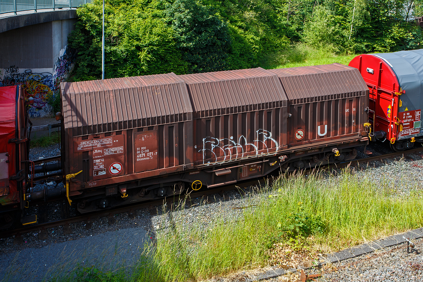 Vierachsiger Drehgestell-Flachwagen mit verschiebbaren Teleskophauben f�r Coiltransporte, 31 80 4671 071-7 D-DB, der Gattung Shimmns-u 708, der DB Cargo AG, am 12 Juni 2024 auf dem KSW Rangierbahnhof Herdorf (Betriebsst�tte Freien Grunder Eisenbahn) , eigereiht im Zugverband. 

Der Wagen eignet sich speziell f�r den Transport witterungsempfindlicher Blechrollen. Er besitzt feste Stirnw�nde, 5 fest im Untergestell eingebaute Lademulden und drei teleskopartig angeordnete Hauben, die sich zu beiden Wagenenden hin soweit durch Ineinanderschieben �ffnen lassen, dass jeweils 2/3 des Wagens freigelegt werden kann. Hierdurch ist eine einfache Beladung des Wagens sowohl von oben als auch von der Seite aus m�glich. Der Wagen darf nur mit geschlossenen und verriegelten Hauben verkehren.

Das Untergestell und die festen Stirnw�nde, die als Abkantrahmenkonstruktion mit Blechbekleidung und hutf�rmigen Verst�rkungsprofilen ausgef�hrt sind, sowie die fest auf den Au�enlangtr�gern angeordneten drei Laufschienen je Wagenseite bilden den als Schwei�konstruktion ausgef�hrten Wagenkasten. Das Untergestell mit seinen beiden kr�ftigen Au�enlangtr�gern besteht aus Walz- und Abkantprofilen. Die Untergestellenden bestehen aus den Hauptquertr�gern, den AK-Tr�gern und den Kopfst�cken. Die sattelf�rmig angeordneten, jeweils mit den Au�enlangtr�gern verschwei�ten Quertr�ger bilden zusammen mit 6 mm dicken Muldenblechen die Lademulden.

Die teleskopartig ineinander verschiebbaren Stahlhauben bestehen aus einer Hohlprofil-Rahmenkonstruktion und einer mit Sicken versehenen Beblechung. Die Abdichtung der Hauben untereinander und zu den Stirnw�nden hin erfolgt �ber labyrinthartig ausgef�hrten Umrandungsprofilen. Die Endhauben verriegeln in geschlossener Stellung automatisch. Das Entriegeln der Hauben ist sowohl vom Erdboden wie auch von der Rampe m�glich. Die beidseitig an jeder Stirnwand angeordneten Verriegelungseinrichtungen sind durch Wellen miteinander verbunden. Hierdurch ist das Entriegeln von nur einer Wagenseite aus m�glich. Jede Haube l�uft auf 4 jeweils und mit Abhebesicherungen versehenen Laufwagen mit nadelgelagerten Doppelrollen.

Die Wagen der Bauart Shimmns 708 sind mit 12 verstellbaren Festlegearmen ausger�stet, die in fest auf den Muldenkappen angeordneten kammartig ausgef�hrten Halterungen gelagert sind. Die Festlegearme und ihre Anordnung ergeben einen wirksamen Schutz gegen unzul�ssiges seitliches Verschieben der Coils.

Um unzul�ssige Radlastabweichungen auszuschlie�en, ist eine m�glichst mittige Verladung der Coils erforderlich. Ein durch alle Mulden in Wagenmitte verlaufender wei�er Farbstreifen dient hierzu als Orientierungshilfe.

TECHNISCHE DATEN:
Erstes Lieferjahr der  �ltesten Wagen:1978
Spurweite: 1.435 mm
Achsanzahl: 4 (in 2 Drehgestelle)
L�nge �ber Puffer: 12.040 mm
L�nge der Ladefl�che: 10.800 mm
Ladebreite (Mulden): 2.400 mm
Drehzapfenabstand: 7.000 mm
Achsabstand im Drehgestell: 1.800 mm
Laufraddurchmesser: 920 mm (neu)
Eigengewicht:  20.810 kg
H�chstgeschwindigkeit: 100  km/h (beladen) / 120 km/h (leer)
Maximales Ladegewicht: 67,0 t (ab Streckenklasse D4) / 61,0 t bei DB ab CM3
Kleinster bef. Gleisbogenradius: 35 m
Bauart der Bremse: KE-GP (LL)
Bremssohle: IB 116
Handbremse: Ja
Intern. Verwendungsf�higkeit: RIV
